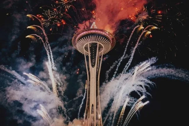 The image features the Space Needle illuminated by colorful fireworks, creating a vibrant and festive atmosphere against a dark night sky.