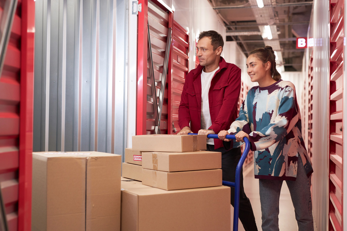 A man and woman are pushing a cart loaded with boxes through a storage facility with red storage units. Both are smiling, engaged in conversation.