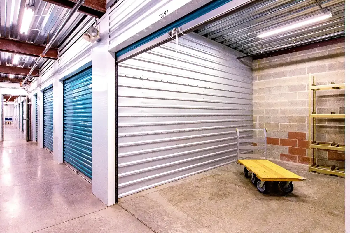 The image shows a storage facility hallway with several storage units, featuring metal doors and a yellow flat cart nearby, all set in a well-lit environment.