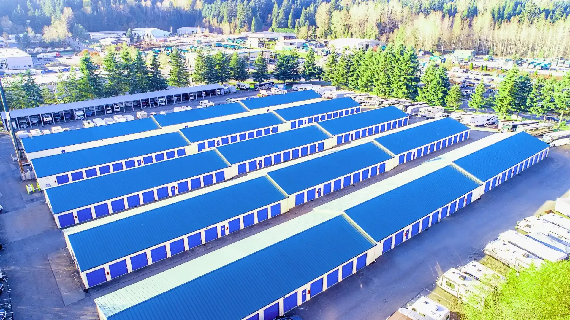 The image shows a large storage facility with multiple units, featuring blue roofs arranged in neat rows, surrounded by trees and open space.