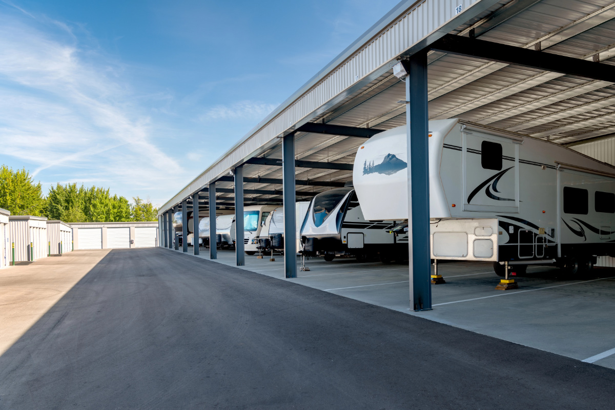 The image shows a row of parked RVs in a shaded storage facility, with clear blue skies in the background and neatly organized spaces.