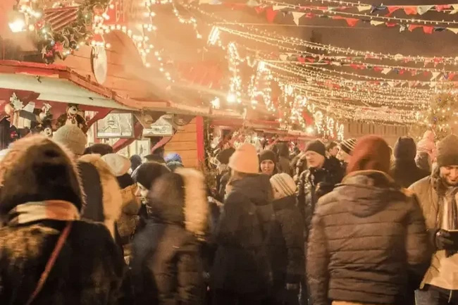 A busy outdoor market filled with people, surrounded by festive lights and decorations, creating a warm and lively atmosphere during the holiday season.