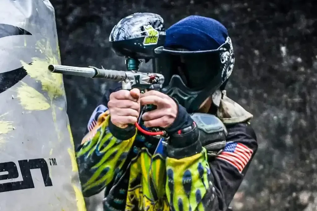 A paintball player in protective gear aims their marker behind a barricade, showcasing intense concentration and readiness in an outdoor arena.